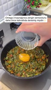 Cracking a fresh egg over the bed of sautéed purslane and onions in a skillet.