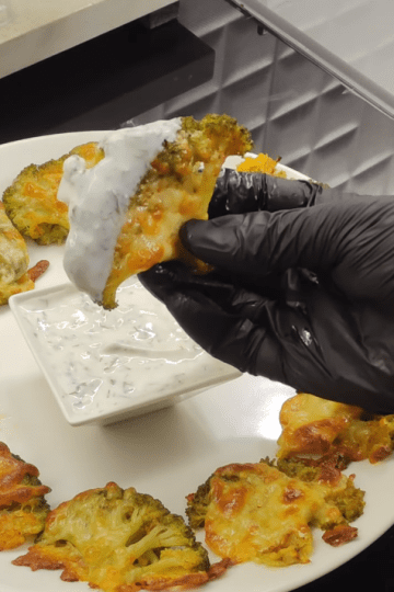 A tray of golden-brown smashed broccoli topped with melted kashar cheese, served with a side of garlic yogurt dip.