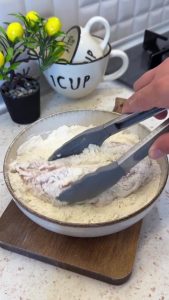 Coating a marinated chicken fillet evenly with all-purpose flour on a plate.