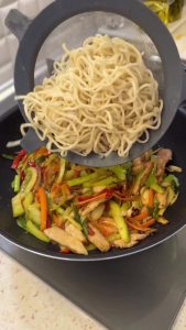 Adding cooked noodles to the pan of sautéed chicken and vegetables.
