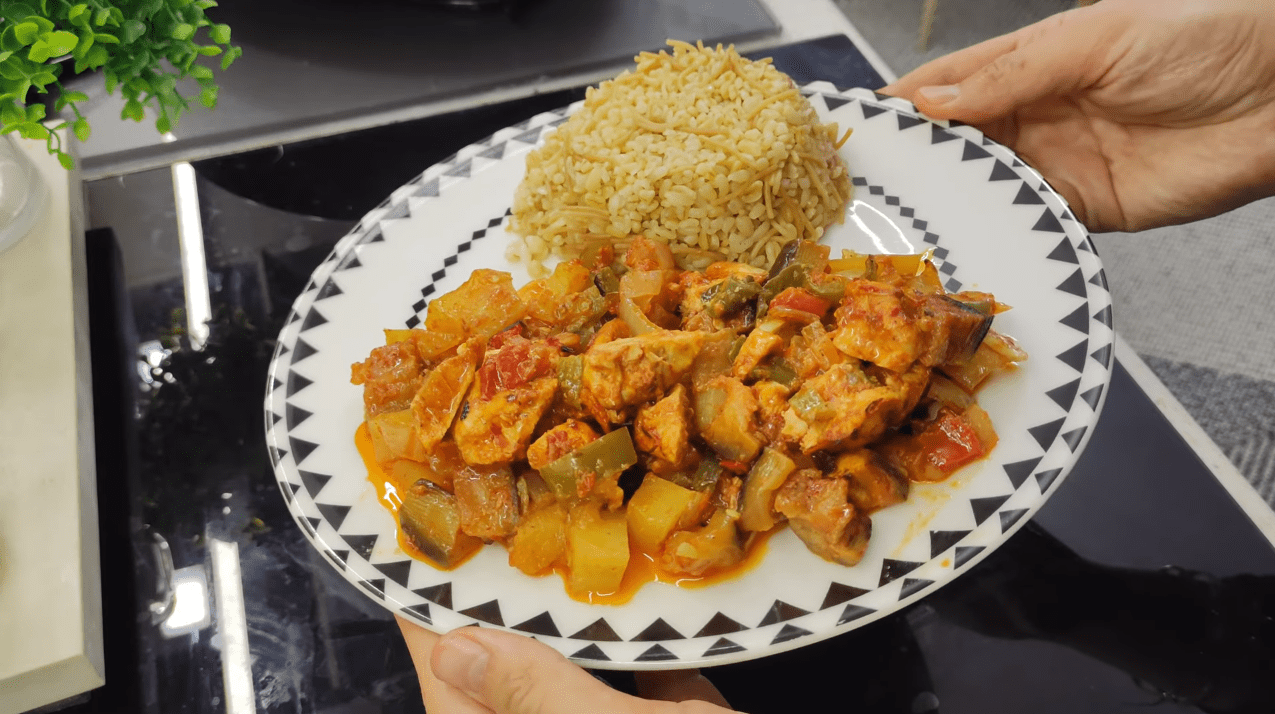 A colorful baking tray filled with roasted chicken cubes, eggplants, potatoes, and peppers in a rich tomato sauce.