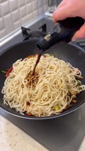 Pouring soy sauce over the noodles and vegetables in a large pan.