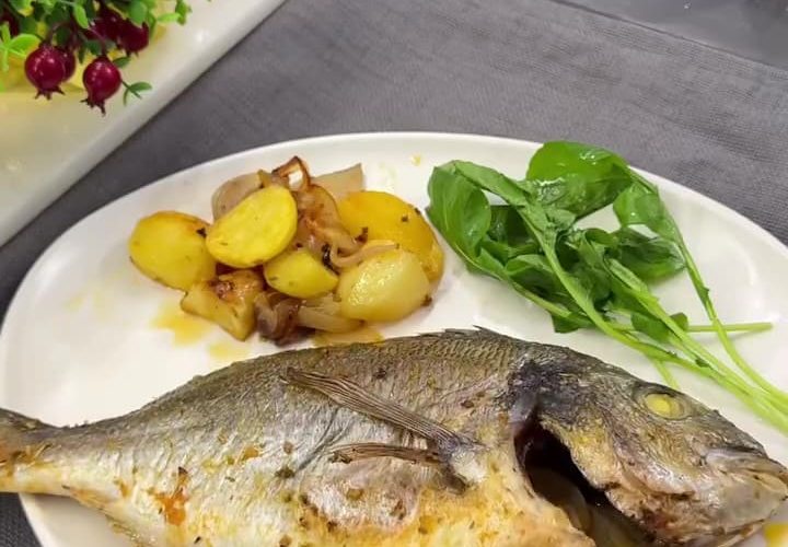 A tray of golden roasted sea bream with baby potatoes and onions, garnished with lemon.