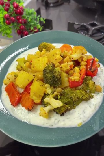 A vibrant platter of roasted spiced winter vegetables including cauliflower, broccoli, and chickpeas, served over a creamy bed of garlic yogurt and fresh dill.