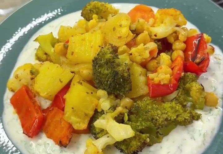 A vibrant platter of roasted spiced winter vegetables including cauliflower, broccoli, and chickpeas, served over a creamy bed of garlic yogurt and fresh dill.