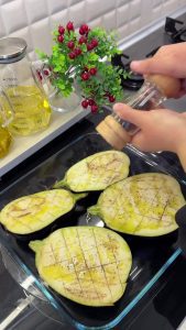 Drizzling olive oil and sprinkling salt and pepper over halved eggplants on a baking tray.