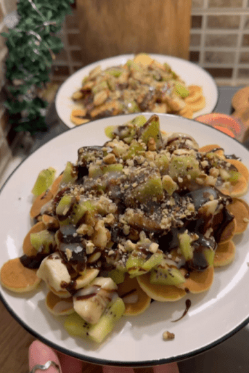 A bowl of golden mini pancake cereal topped with sliced kiwi, banana, walnuts, and chocolate sauce.