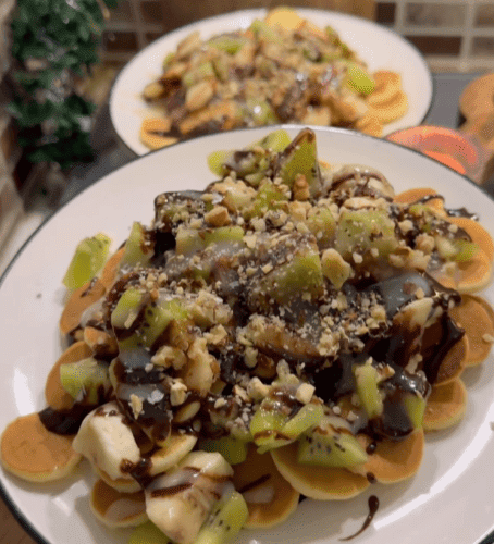 A bowl of golden mini pancake cereal topped with sliced kiwi, banana, walnuts, and chocolate sauce.