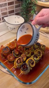 Pouring a red tomato and pepper paste sauce over the eggplant rolls in a tray.