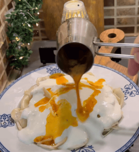 A plate of Turkish mantı dumplings topped with white garlic yogurt and red spicy butter sauce.