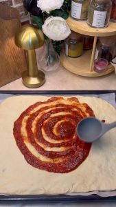 Spreading a zesty tomato and oregano sauce over the raw pizza dough in the tray.
