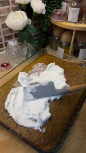 Spreading a smooth layer of white whipped cream over the soaked revani cake.
