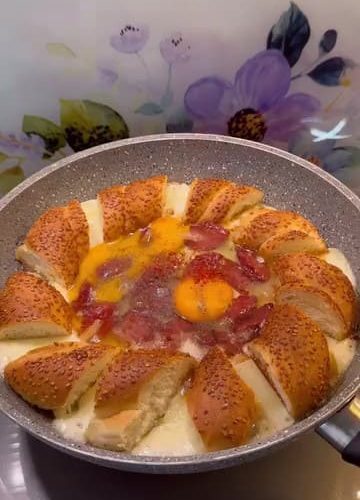 A sizzling breakfast skillet with simit pieces, sucuk, melted cheese, and eggs.